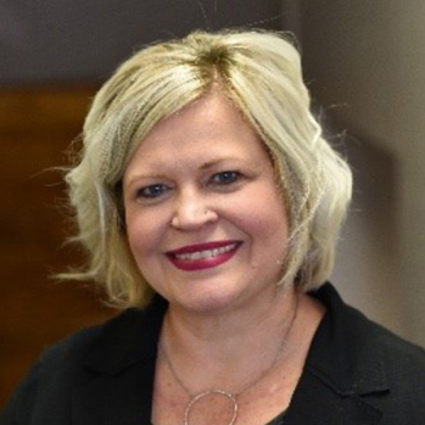 Head and shoulders of smiling Dawn Mandt, short blonde wavy hair, light pale skin, dark suit jacket on blurry background