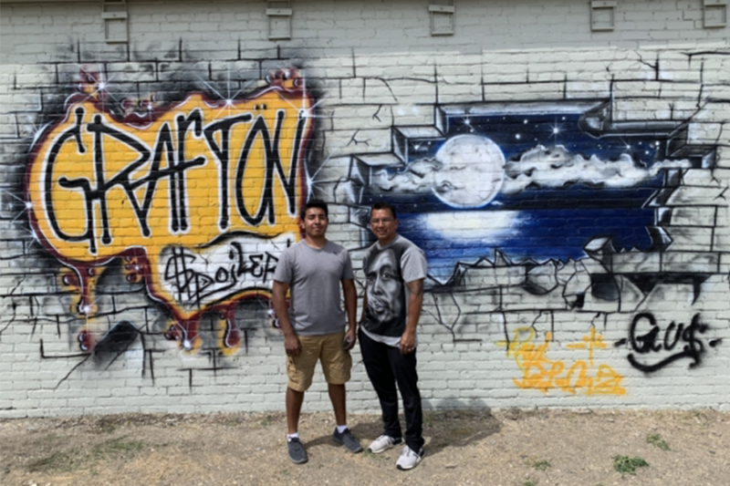 2 people standing in front of brick wall, turned graffiti mural with Grafton spelled out