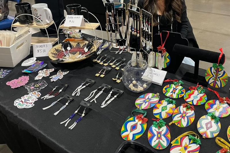 Sacred Pipe Resource Center Art Fair showing a black table with various Indigenous colorful artworks, including mugs, jewelry and holiday ornaments