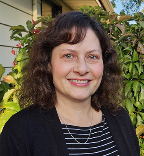 Head and shoulders of Lisa McCallum with fair skin, hazel eyes, shoulder-length curly brown hair wearing a black shirt with thin white lines and a black sweater, standing outside in front of leafy green bushes