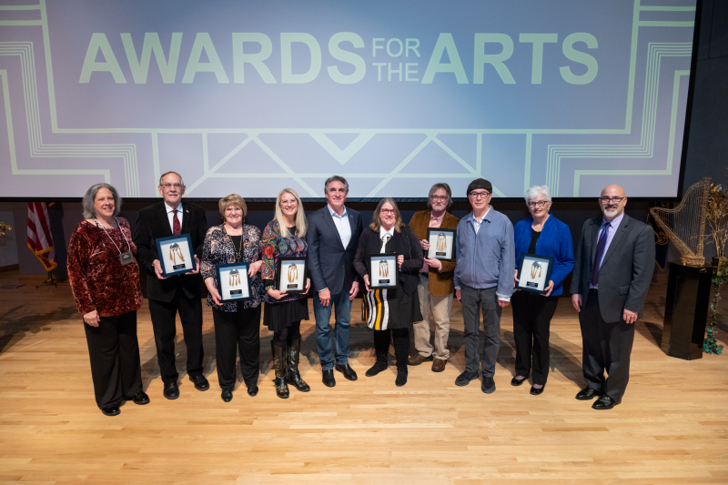 Ten adults standing on a hard wood floor, dressed business casual, holding six large black-framed awards with a large projected screen in the background that reads AWARDS FOR THE ARTS. 