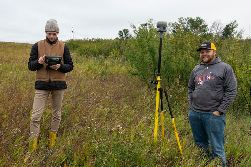 Two men standing outside on an overgrown field on a fall day with surveying equipment