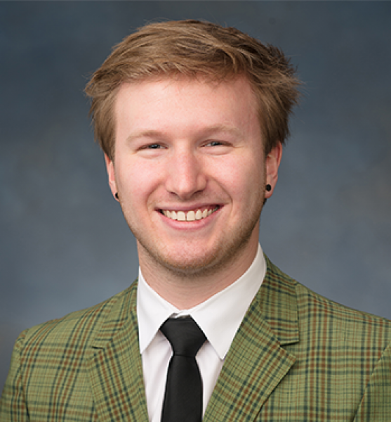 head and shoulders of Tobias Zikmund with short, messy, dirty blonde hair, black dot earrings, pale skin and a big toothy smile; wearing a green striped suit jacket and black tie