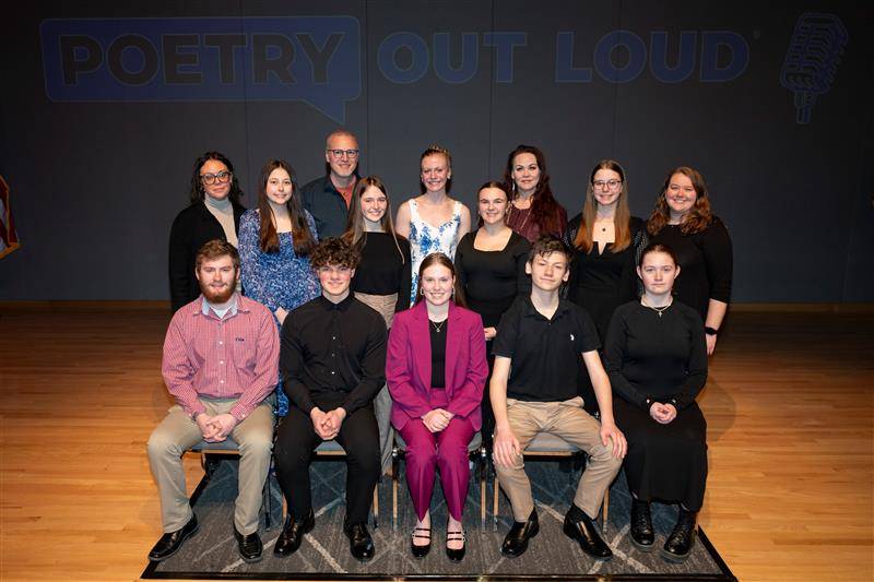 Three rows showing ten high school Poetry Out Loud school champions, along with four adults who coordinated the event, standing on stage during the ND POL State Championship. The words Poetry Out Loud are projected on the wall behind them. 