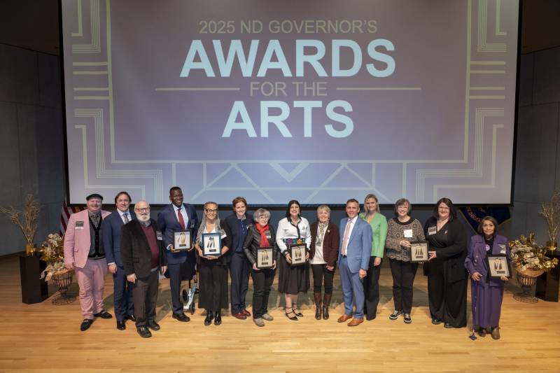 Large group photo with ND Governor's Awards for the Arts on screen above a stage with all seven recipients and ND Governor and First Lady Armstrong
