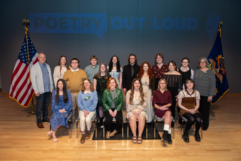Two rows showing sixteen high school Poetry Out Loud champions in the middle and an adult male and adult female, who work for ND Council on the arts, standing on the ends. The words Poetry Out Loud are projected on the wall behind them. There is a USA flag and North Dakota flag on both ends as well.